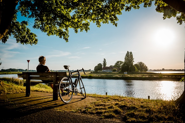 Radfahrer ruht sich auf einer Bank an der Maas aus und genießt den Sonnenuntergang in Stevensweert